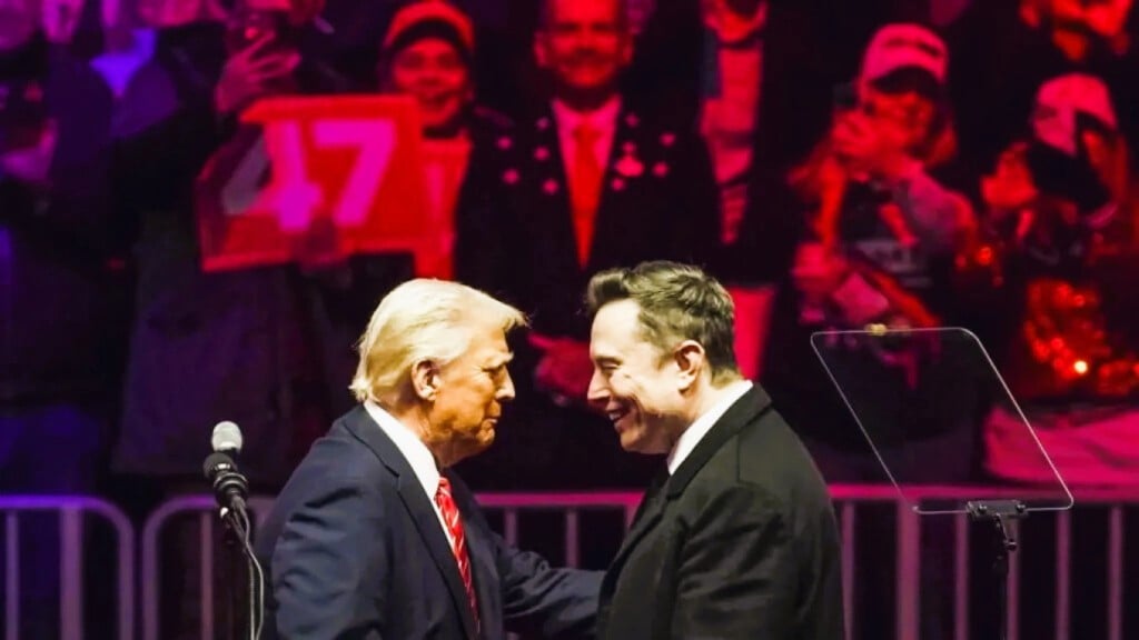 Donald Trump and Elon Musk shake hands at a public event, with supporters in the background.