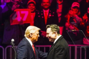 Donald Trump and Elon Musk shake hands at a public event, with supporters in the background.